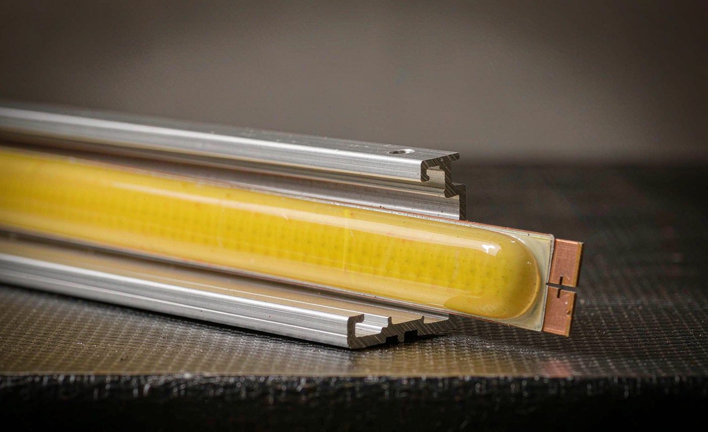 Macro photograph of a COB LED strip: one continuous yellow phosphor coating over the full length, seated in an aluminium profile with a visible solder pad and cut mark
