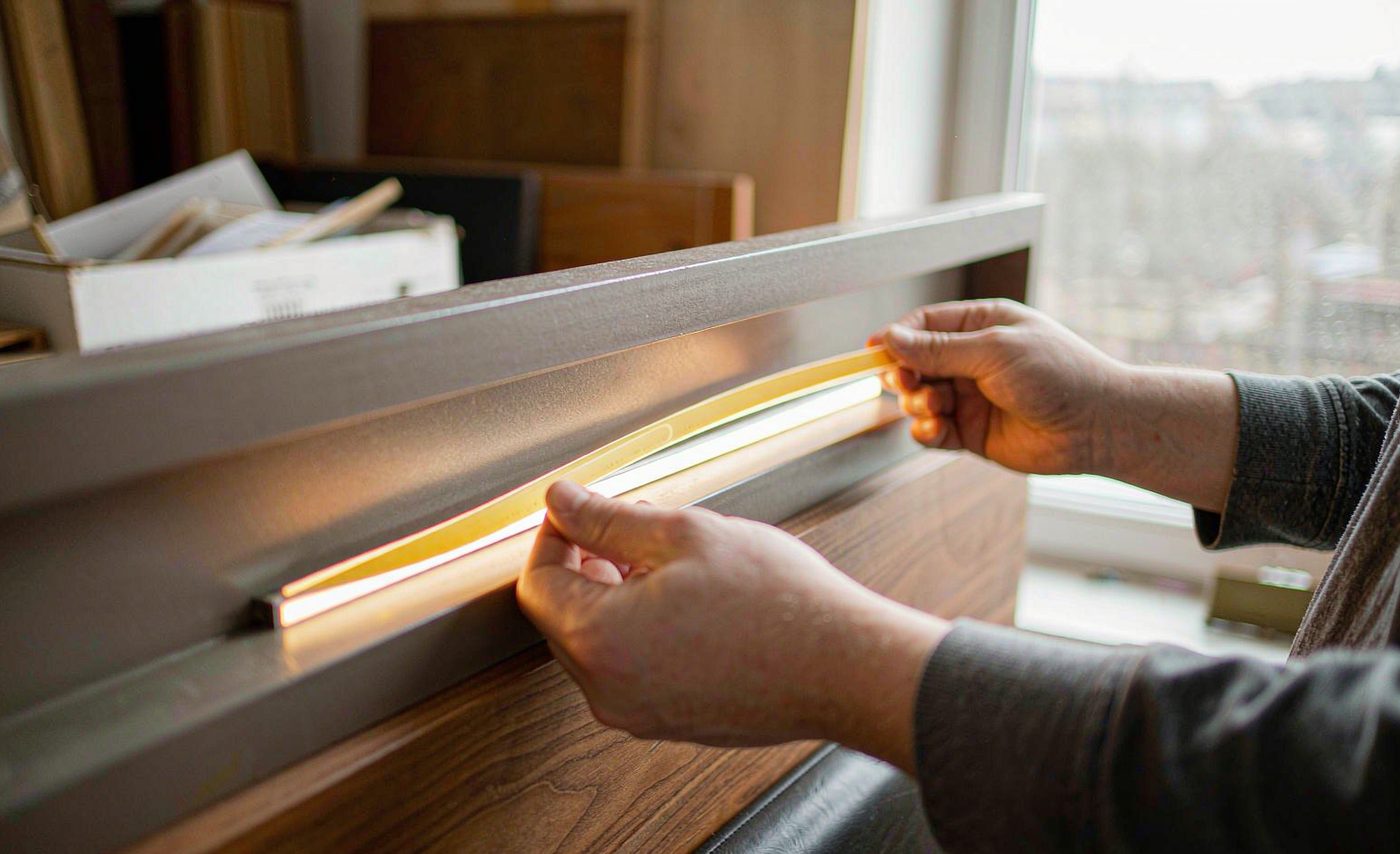A technician seating a flexible warm-white COB LED strip into an aluminium channel recessed into a wooden headboard; the lit strip is one continuous unbroken line of warm light